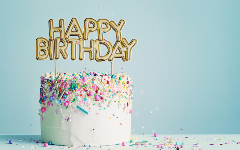 Gâteau d'anniversaire blanc posé sur une table blanche avec des confettis d'anniversaire et une bougie "joyeux anniversaire" posé sur le gâteau . il y a un fond bleu