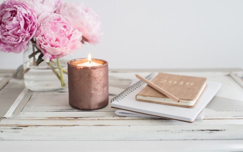 Bougie parfumée allumée posée sur une table en bois, à côté d’un carnet et de fleurs roses, créant une ambiance douce et inspirante propice à la détente.