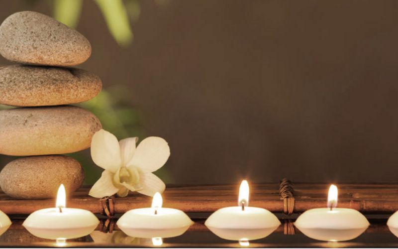 Bougies flottante allumées alignées devant des galets empilés et une fleur blanche, créant une ambiance zen et apaisante.