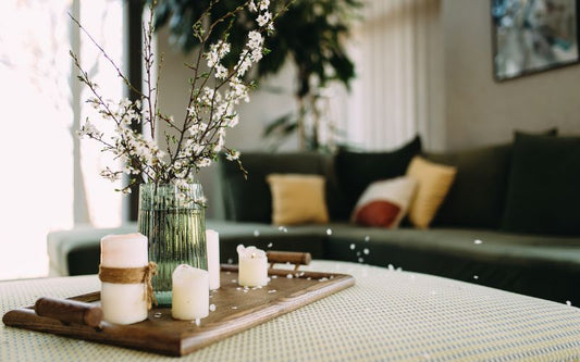 Un plateau de bougie est disposé sur une table basse de salon avec des fleurs à côté, on peut y voir un canapé derrière. Ambiance cocooning..