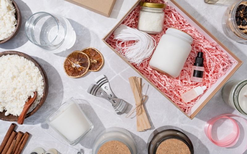 Il est disposé sur une table blanche, des bougies et du matériel de fabrication de bougie ainsi qu'un coffret cadeau.