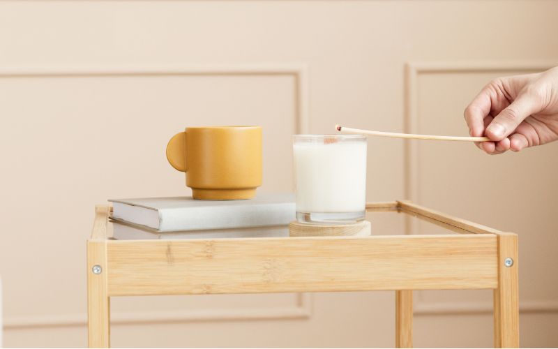 Une personne est en train d'allumer une bougie blanche dans un contenant en verre, elle est posée sur une table à côté d'un livre, Ambiance zen. 