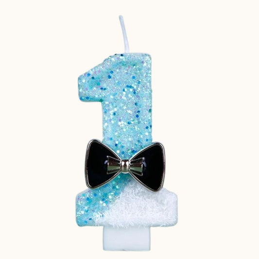 Bougie Anniversaire Charme Scintillant, le chiffre représenté est le 1, de couleur bleue et blanche avec un nœud noir. Sur fond beige clair.