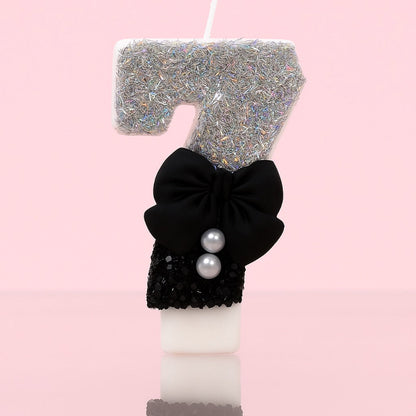 Bougie Anniversaire Nœud Chic, le chiffre représenté est le 7, de couleur noire et grise, avec un noeud et des paillettes. Sur fond rose pastel.
