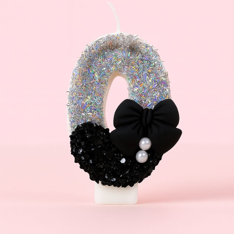 Bougie Anniversaire Nœud Chic, le chiffre représenté est le 0, de couleur noire et grise, avec un noeud et des paillettes. Sur fond rose pastel.