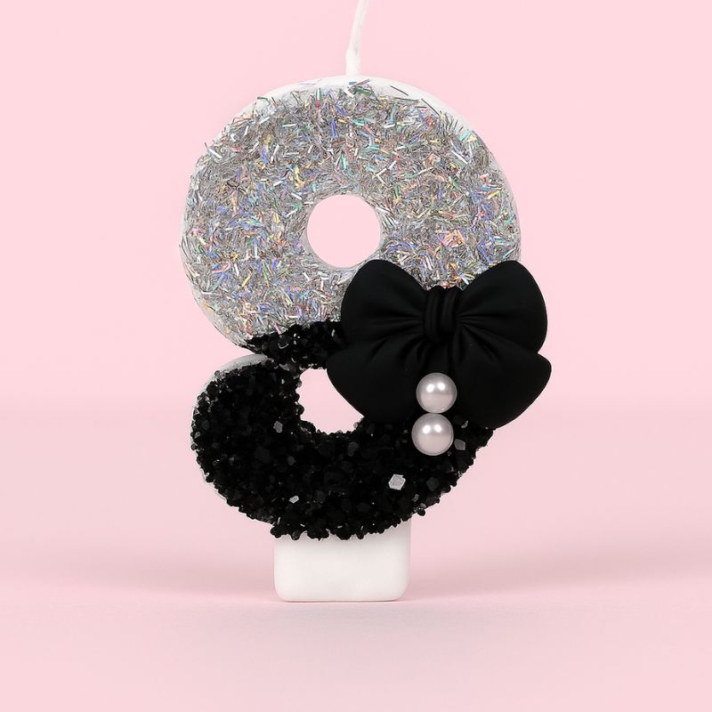 Bougie Anniversaire Nœud Chic, le chiffre représenté est le 9, de couleur noire et grise, avec un noeud et des paillettes. Sur fond rose pastel.