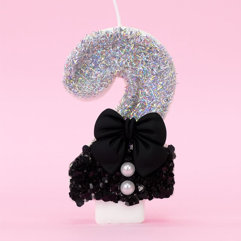 Bougie Anniversaire Nœud Chic, le chiffre représenté est le 2, de couleur noire et grise, avec un noeud et des paillettes. Sur fond rose pastel.