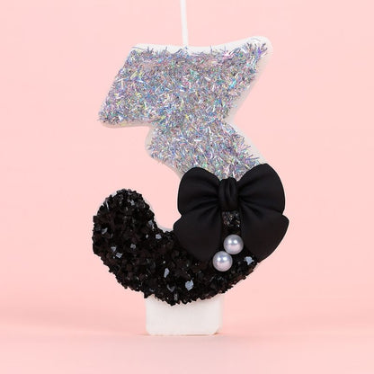 Bougie Anniversaire Nœud Chic, le chiffre représenté est le 3, de couleur noire et grise, avec un noeud et des paillettes. Sur fond rose pastel.