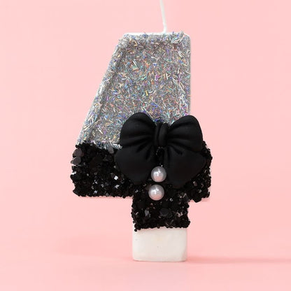 Bougie Anniversaire Nœud Chic, le chiffre représenté est le 4, de couleur noire et grise, avec un noeud et des paillettes. Sur fond rose pastel.
