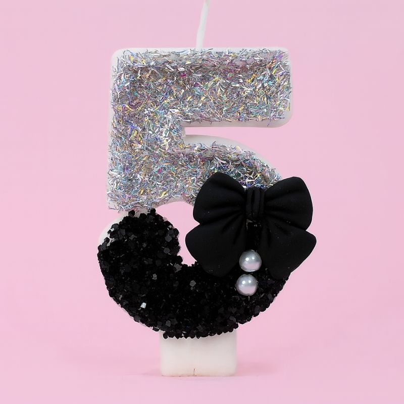 Bougie Anniversaire Nœud Chic, le chiffre représenté est le 5, de couleur noire et grise, avec un noeud et des paillettes. Sur fond rose pastel.