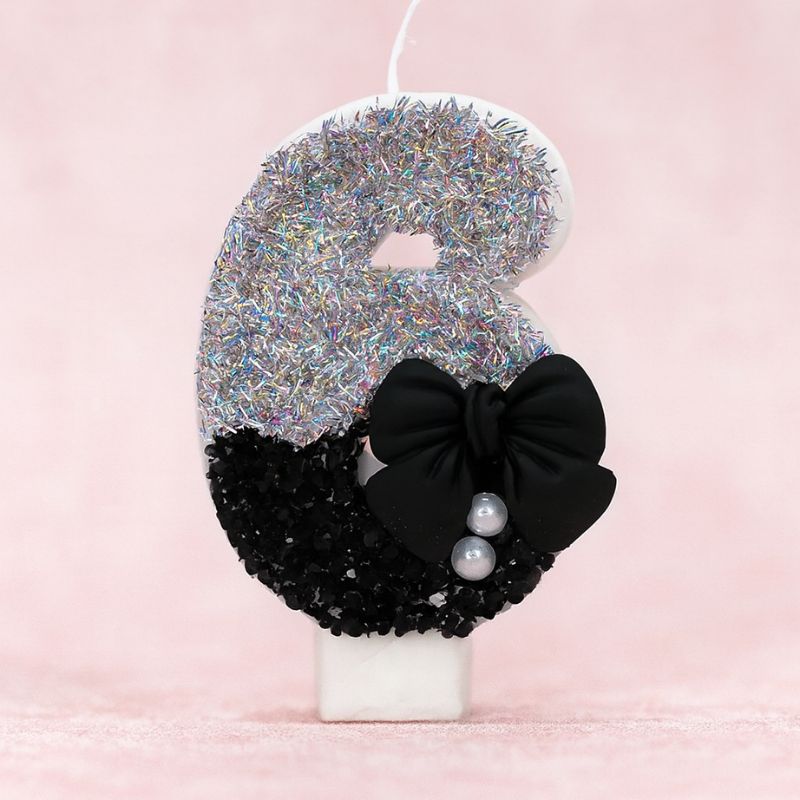 Bougie Anniversaire Nœud Chic, le chiffre représenté est le 6, de couleur noire et grise, avec un noeud et des paillettes. Sur fond rose pastel.