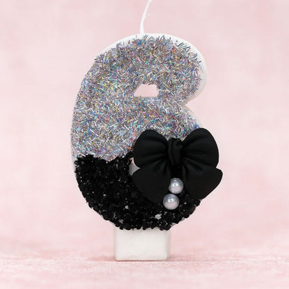 Bougie Anniversaire Nœud Chic, le chiffre représenté est le 6, de couleur noire et grise, avec un noeud et des paillettes. Sur fond rose pastel.