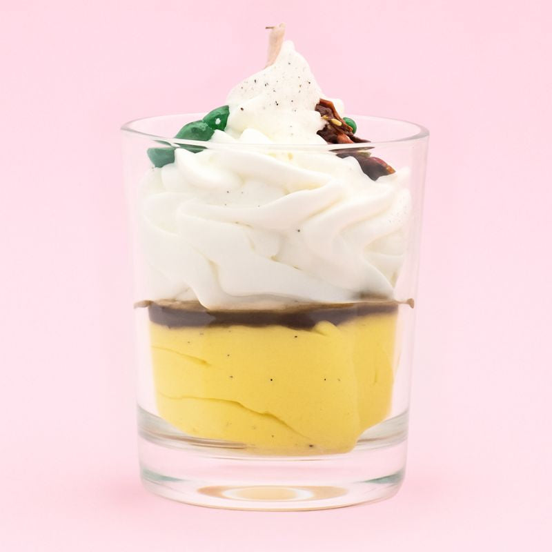Bougie Bijou Surprise Précieuse, de couleur jaune et blanche, avec des petits motifs de Noël sur le dessus de la chantilly. Sur fond rose pâle.