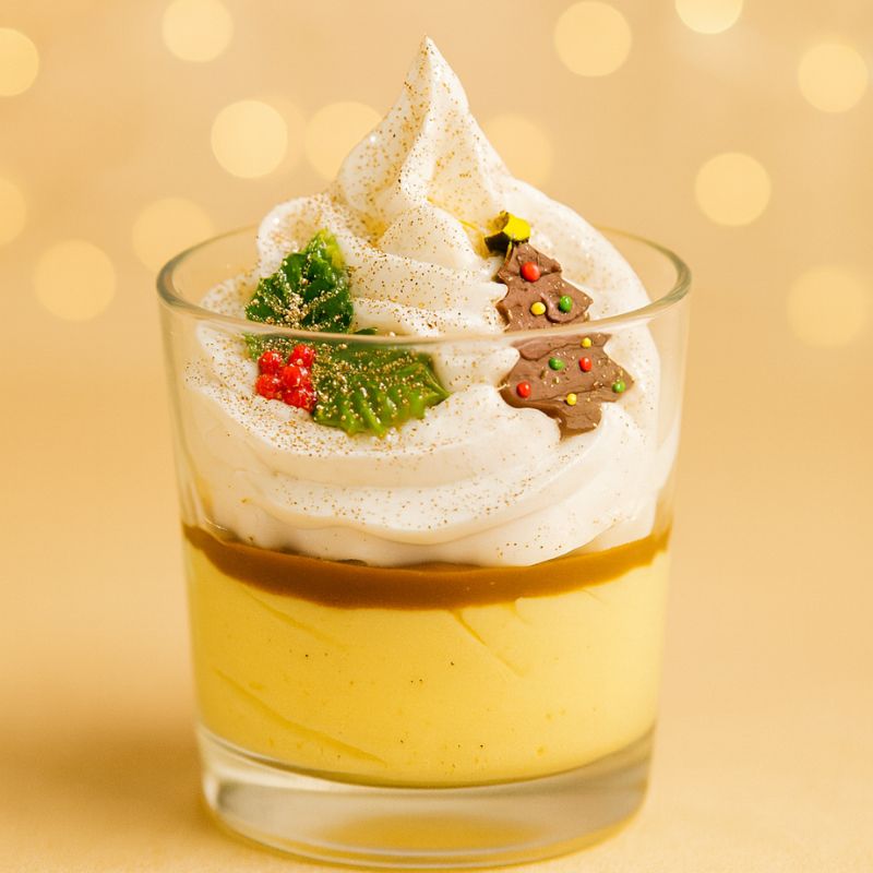 Bougie Bijou Surprise Précieuse, on peut voir une feuille de houx et un sapin sur la chantilly de la bougie bijou. Sur fond jaune doré.