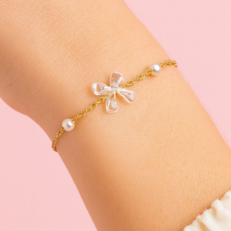 Bougie Bijou Surprise Précieuse, en détail le bijou posé sur un poignet, il est de couleur or, avec un nœud transparent et des perles. Sur fond rose pâle.