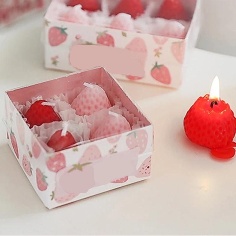 Bougie Décorative Fruit Fraise, dans sa boite de protection, une bougie allumée à côté, le tout posé sur une table.
