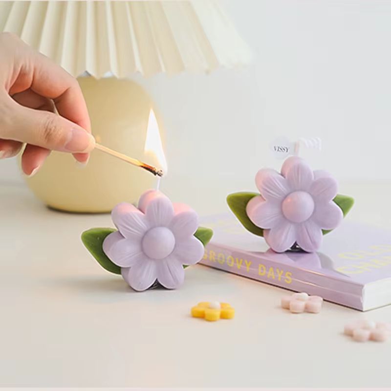 Bougie Décorative Pâquerette, disposée sur une table, il y a un livre et une lampe. La bougie est en train d’être allumée par quelqu’un.