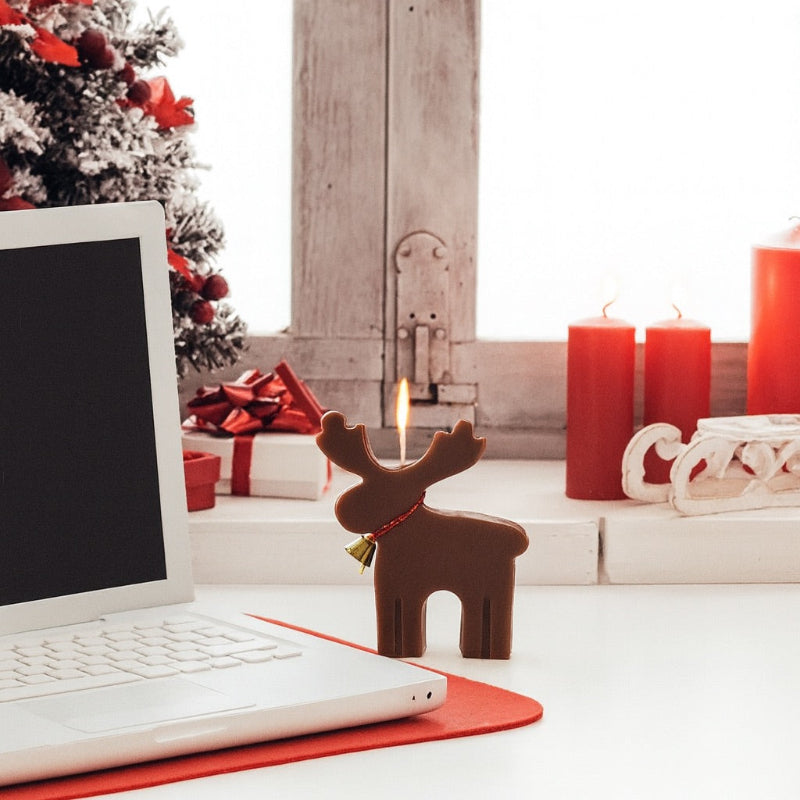 Bougie Noel Féérie du Renne Enchanté, de couleur marron et allumée. La bougie est posée sur un bureau blanc.
