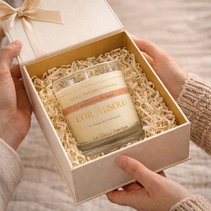 Bougie Parfumée Artisanale L’Or Absolu présentée dans un coffret cadeau élégant, nichée dans un écrin beige avec ruban, mise en scène chaleureuse et raffinée idéale pour offrir un moment précieux.