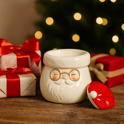 Brule Parfum Magie de Noël, le couvercle du bruleur est posé à côté de celui-ci, il y a des cadeaux autour et le tout est posé sur une table en bois. Esprit de Noël.