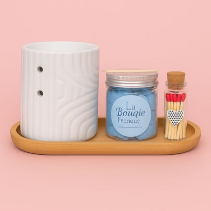 Coffret Bougie Évasion Parfumée, composé d'un brûleur blanc, d'un fondant crémeux au parfum adoucissant, d'allumette et d'un plateau en bois sur un fond rose clair.
