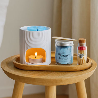 Coffret Bougie Évasion Parfumée, le bruleur est allumé, avec du fondant crémeux à l’intérieur et tous les éléments qui le composent son posé sur un tabouret dans un salon.