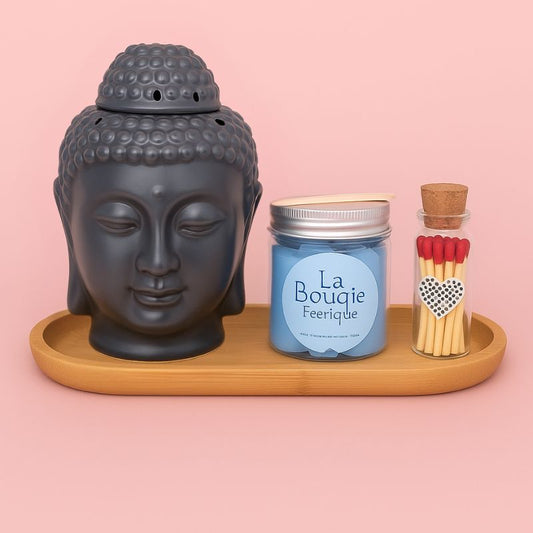 Coffret Bougie Magie des Senteurs, avec un brûleur Bouddha de couleur noir, un fondant parfum adoucissant, des allumettes et un plateau en bois. Sur fond rose clair.