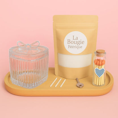 Coffret Bougies Perles Rituel Cocooning, avec un contenant en verre, un sachet de cire perles, des mèches, un plateau en bois et des allumettes, le tout sur un fond rose pâle.
