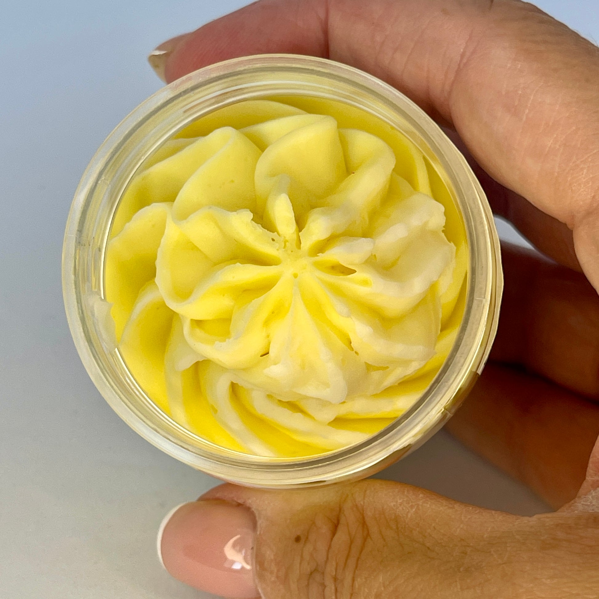 Fondant Parfumé Fleur de Tiaré Envoûtante, fondant dans son contenant, de couleur jaune et tenue dans une main pour présenter. Fond gris.