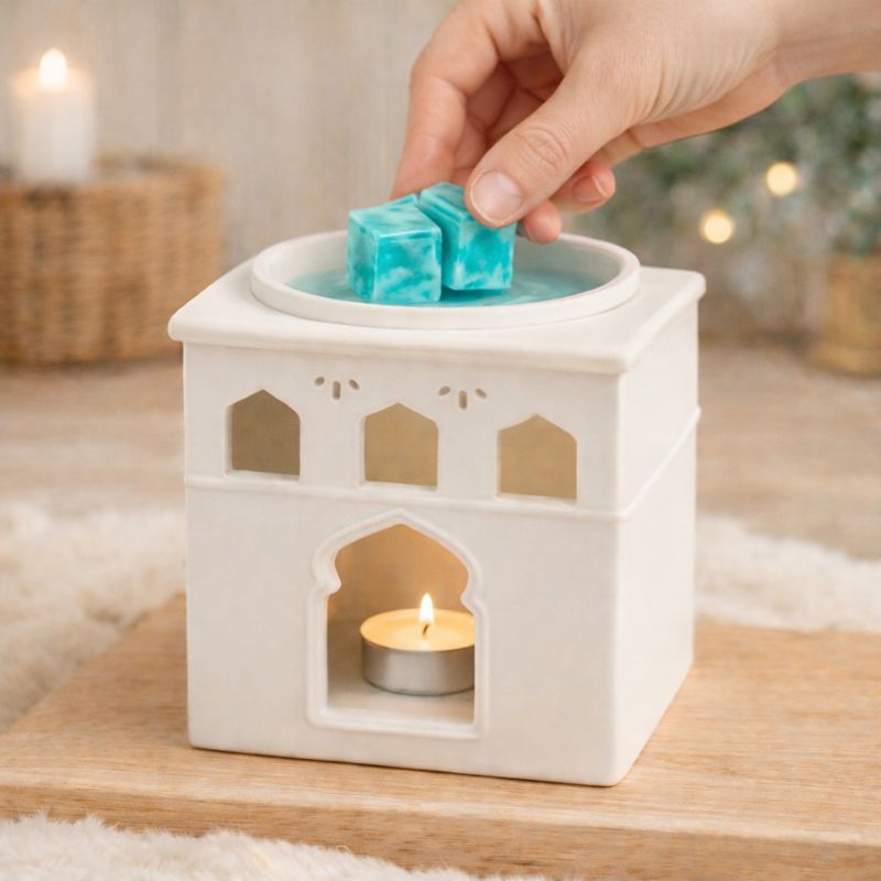 Fondant Parfumé Tablette Chanvre, Main déposant deux carrés de fondant parfumé chanvre turquoise dans un brûle-parfum blanc en céramique avec bougie chauffe-plat allumée, ambiance scandinave chaleureuse.