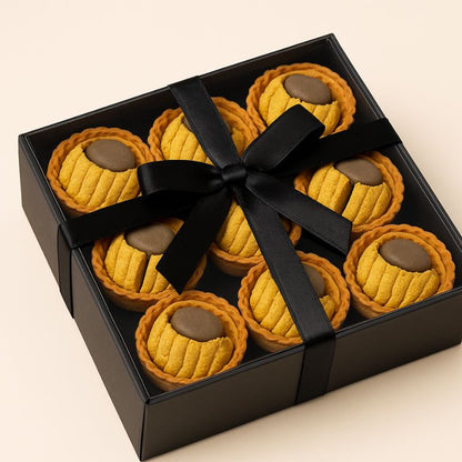 Fondant Parfumé Tartelette Châtaigne Grillé, présenté dans un coffret-cadeau noir avec un noeud. Sur fond beige.