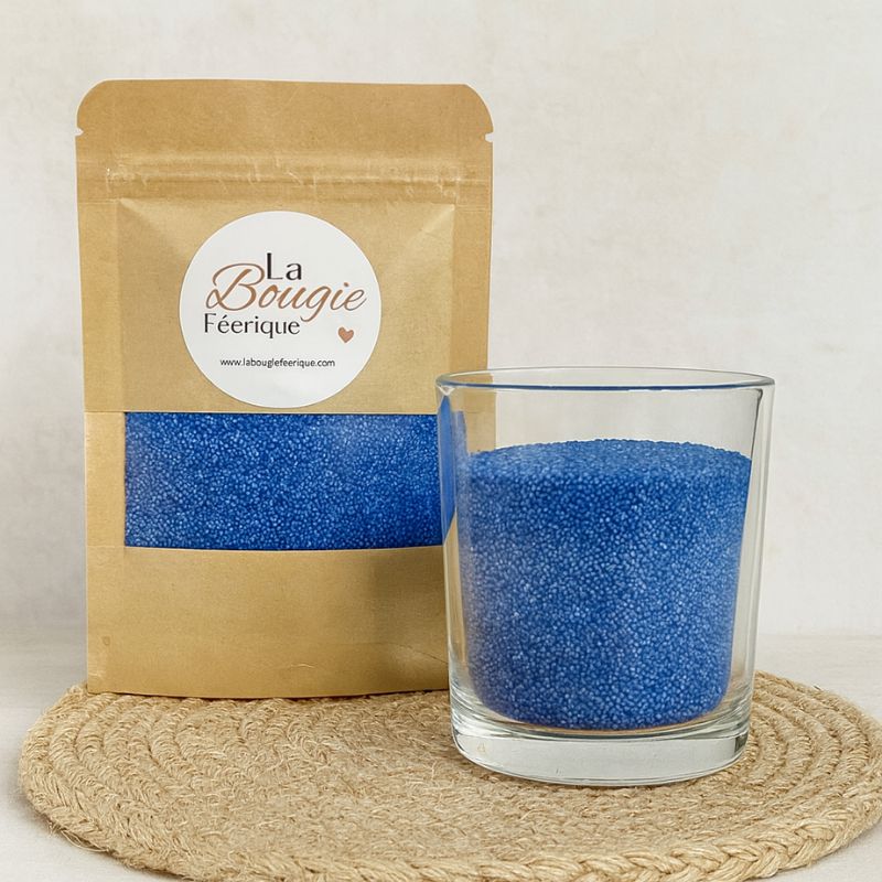 Perles de Cire Décorative pour Bougie, de couleur bleue, avec son contenant transparent où il y a de la cire perle en démonstration. Fond beige.