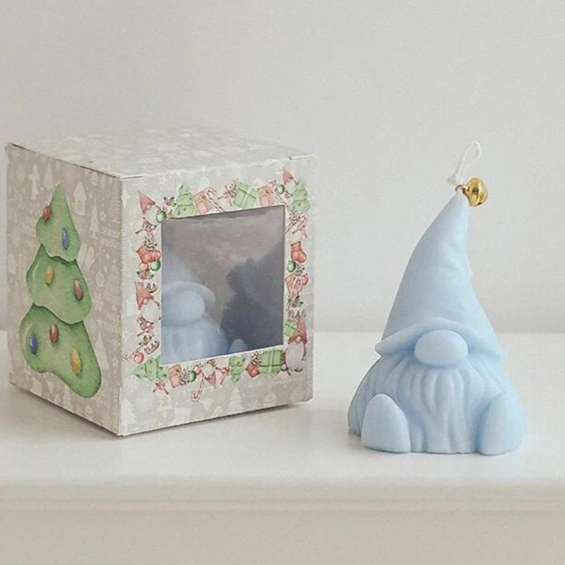 Bougie Décorative Gnome, de couleur bleue et disposée à côté de sa boîte de transport sur ne table. Fond beige clair. 